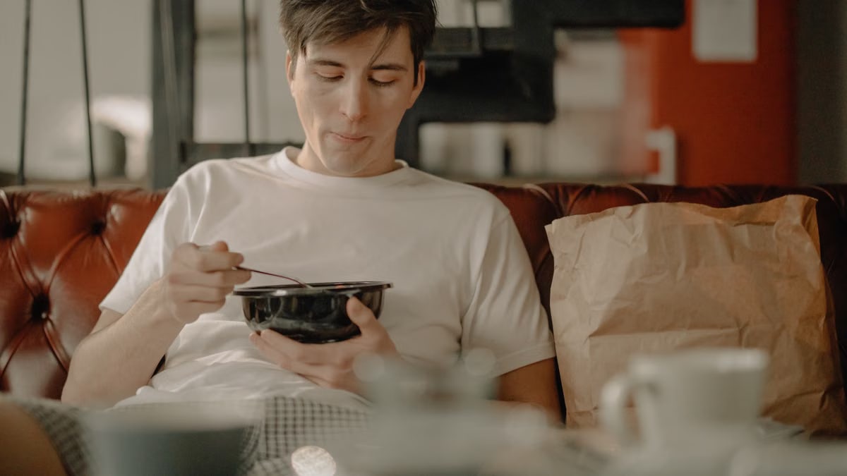 A young man relaxes at home, enjoying a meal with a takeout bag on the couch.
